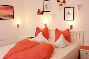 Una cama bien hecha con ropa de cama naranja y blanca, un cabecero blanco y una mesita de noche con una lámpara. Hay estantes montados en la pared con objetos decorativos e imágenes enmarcadas.