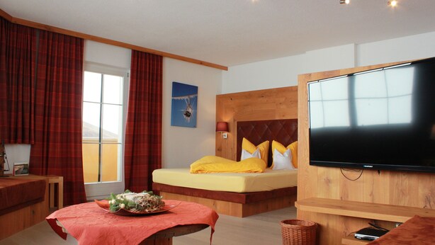 A modern hotel room with a large bed, a flat-screen TV, a wooden headboard, and a window with curtains. - Summer dream vacation apartment in the Zillertal with magnificent views (Schwendau)
