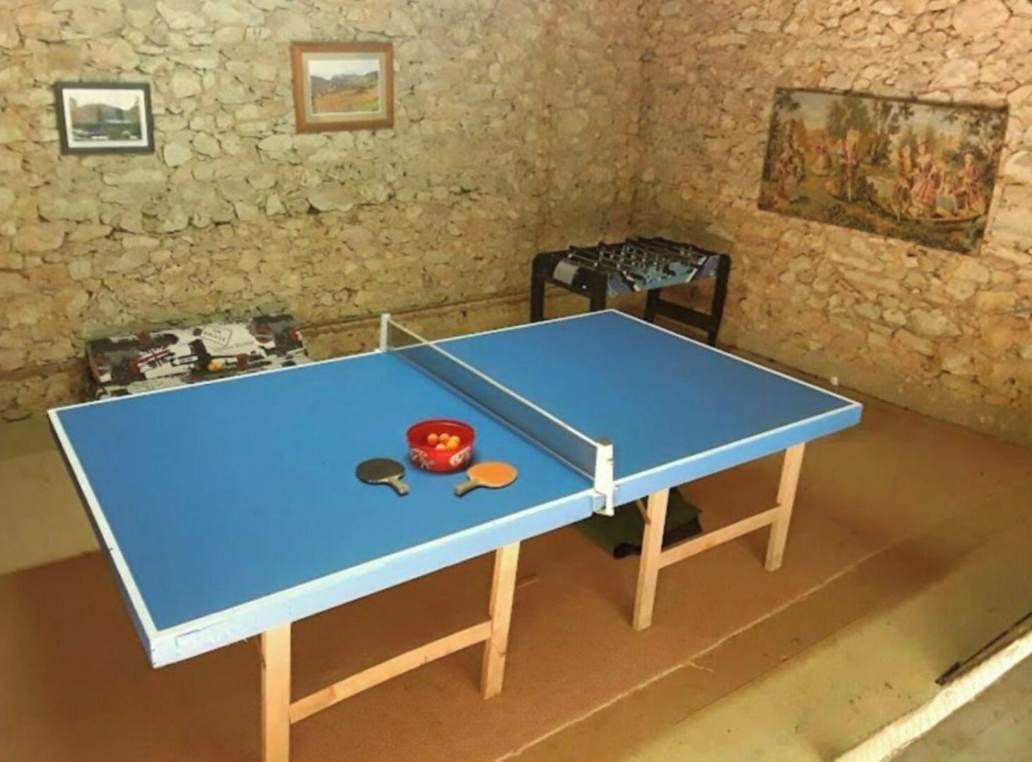 Una mesa de ping pong con dos paletas y un organizador de pelotas en una habitación con paredes de piedra y un cuadro.