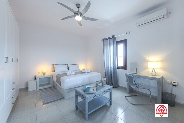 A modern hotel room with a bed, bedside tables, a desk, a chair, a ceiling fan, and an air conditioning unit. - Villa Naya (Santorini)