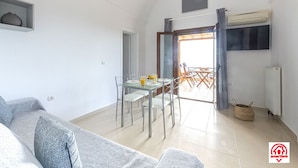 A modern living room with a grey sofa, a dining table with chairs, and a TV mounted on the wall. - Villa Naya (Santorini)