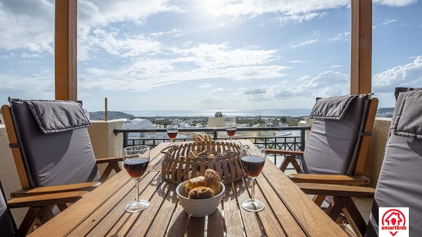 A wooden table with a view of the city, two chairs, a bowl of bread, and two glasses of red wine. - Villa Naya (Santorini)