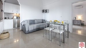 A modern kitchen with white cabinets, a grey sofa, and a dining table with chairs. - Villa Naya (Santorini)