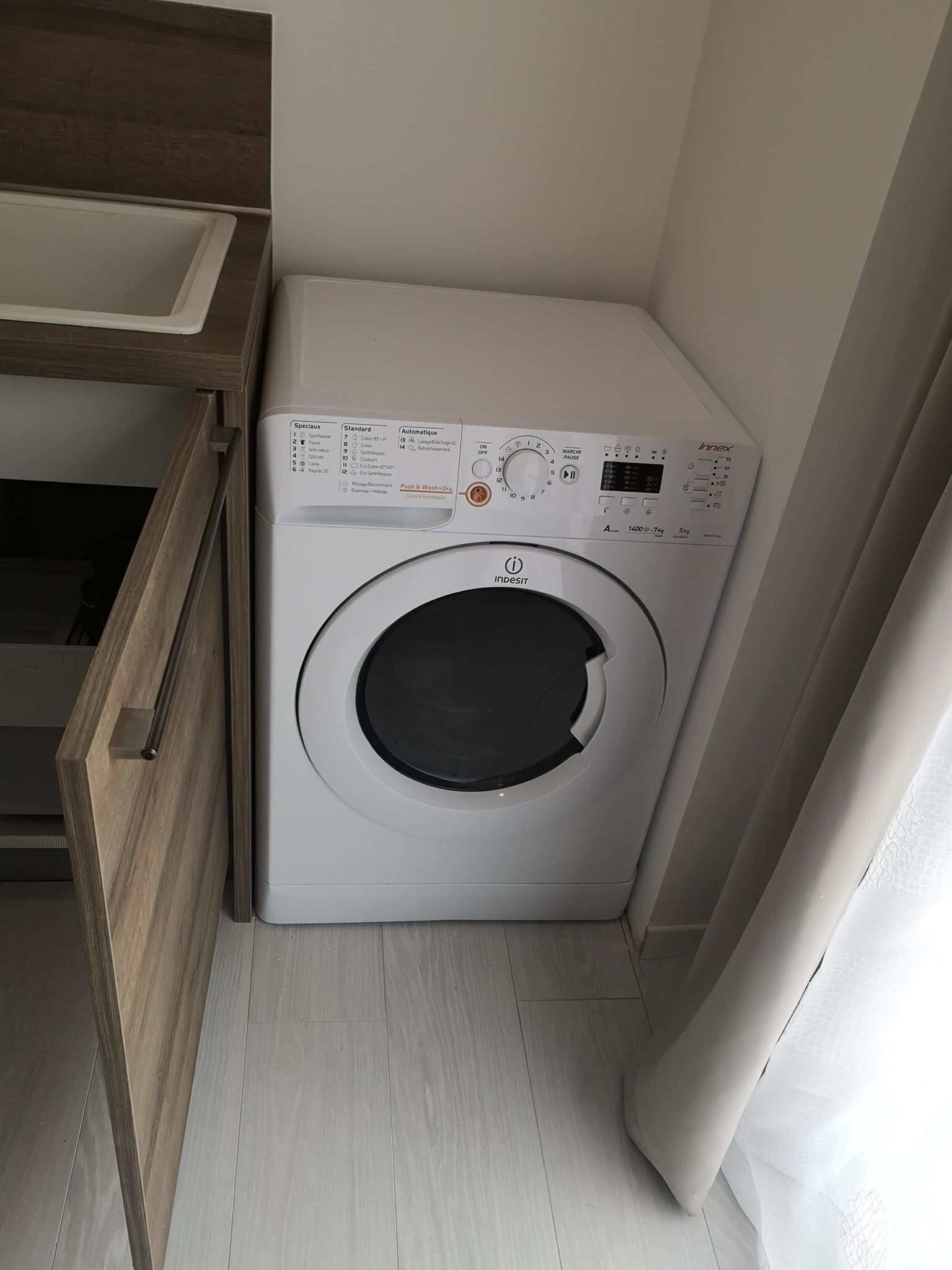 Máquina de lavar roupa branca e compacta com painel de comandos, numa pequena lavanderia com armário de madeira e chão branco.