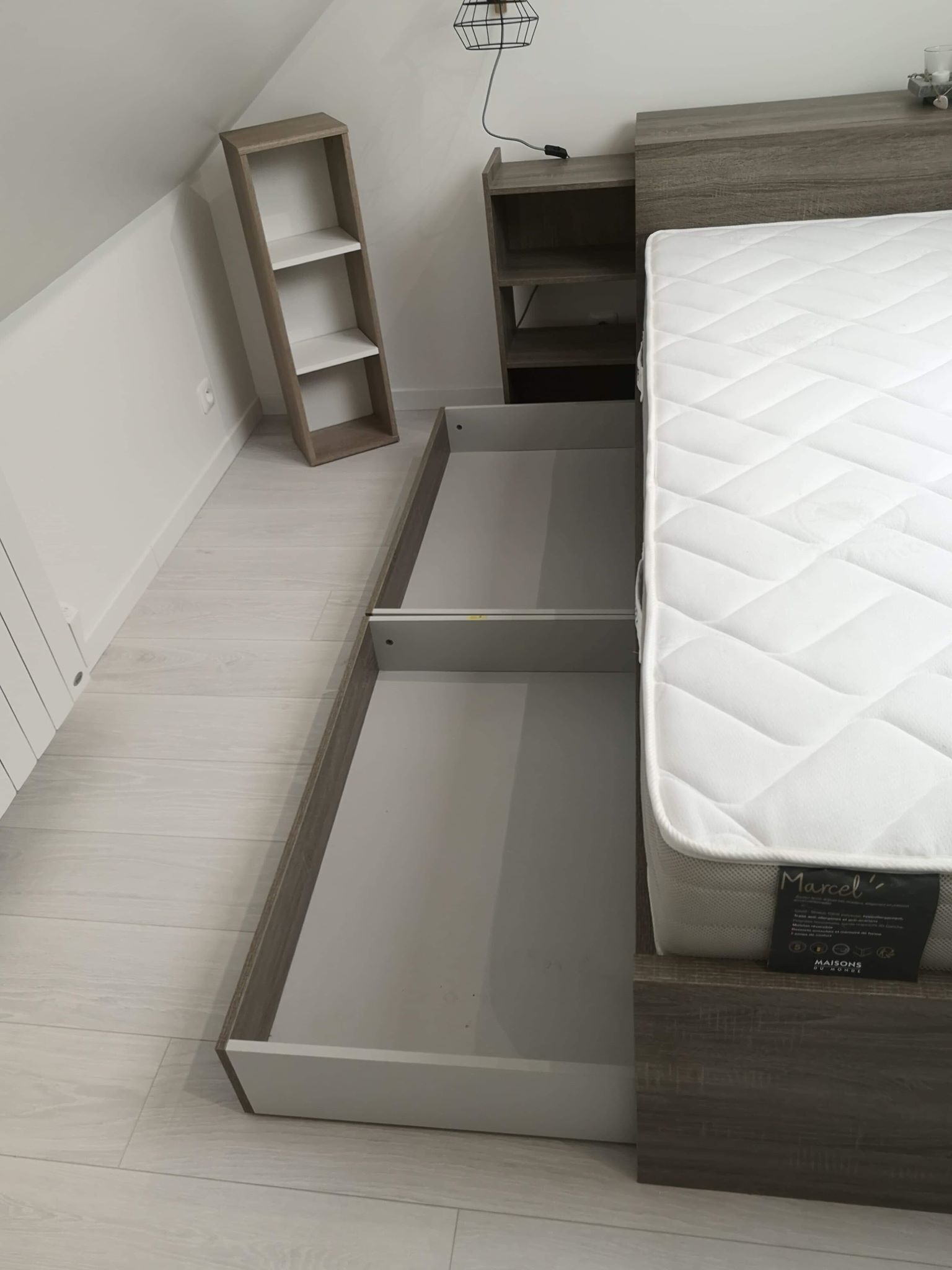 A bedroom with a bed, a wooden nightstand, a small shelf, and a wall-mounted light fixture.