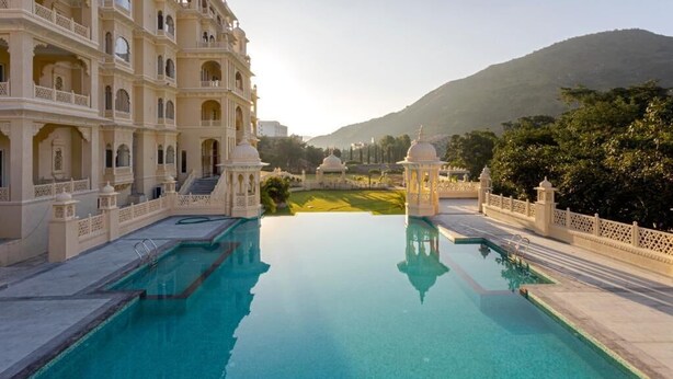 A pool with a reflection of a multi-story building with balconies and a dome structure in the background. - Rawla Sagrun (Kumbhalgarh)