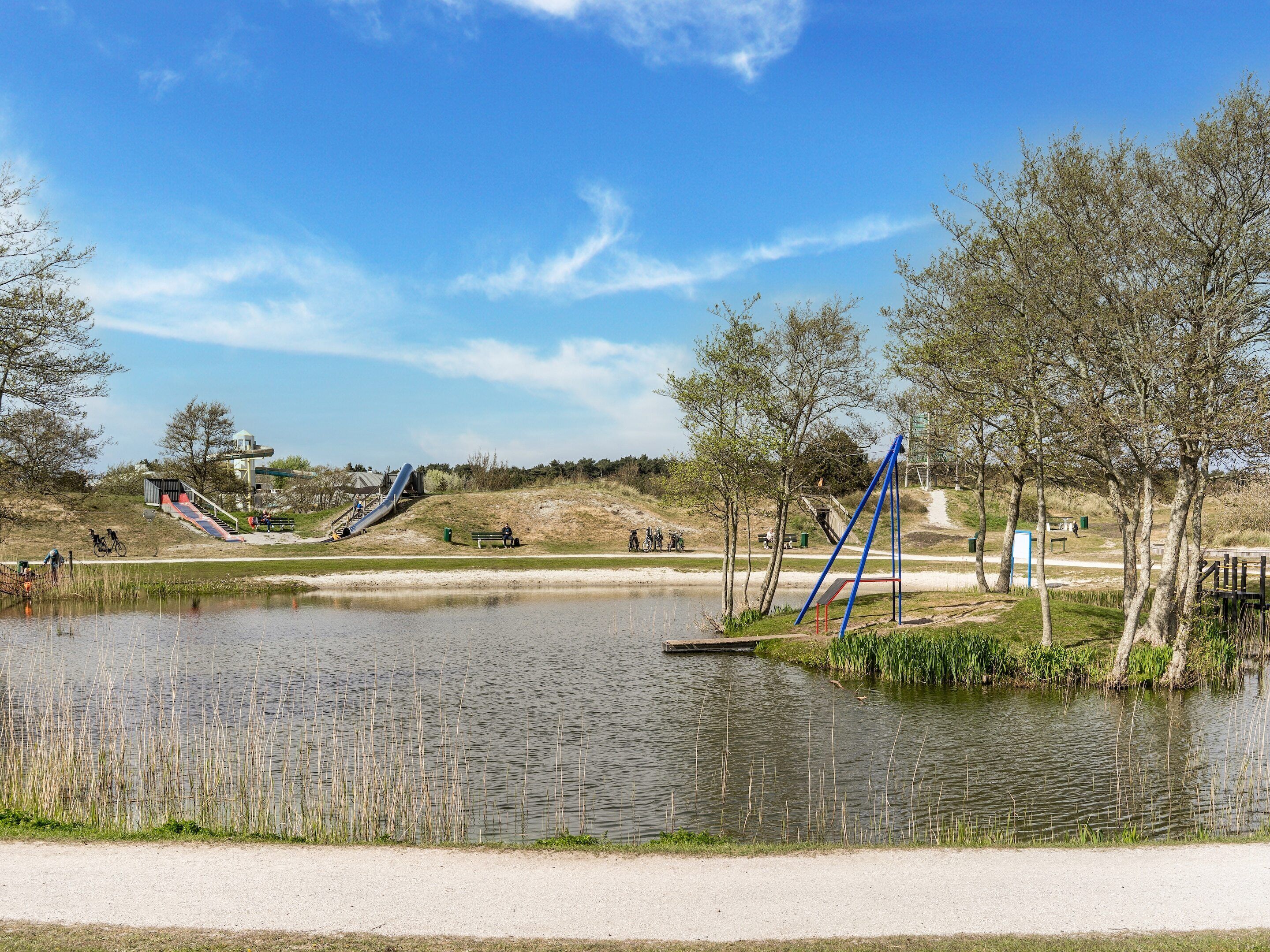 Un parque con un estanque, equipo de juegos infantiles y un sendero de tierra.
