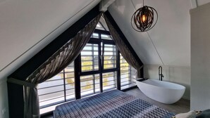 A bedroom with a sloped ceiling, a large window, a white bathtub, and a patterned bedspread.