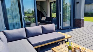 A modern outdoor seating area with a sofa and a table set for a meal, featuring a bottle and glasses. - Water Villa in Balk With Lake Access (Balk)