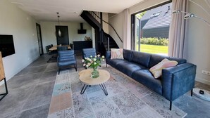 A modern living room with a dark blue sofa, a round coffee table with a vase of flowers, and a patterned rug. There is a staircase leading to an upper level, and a large window with curtains. - Water Villa in Balk With Lake Access (Balk)