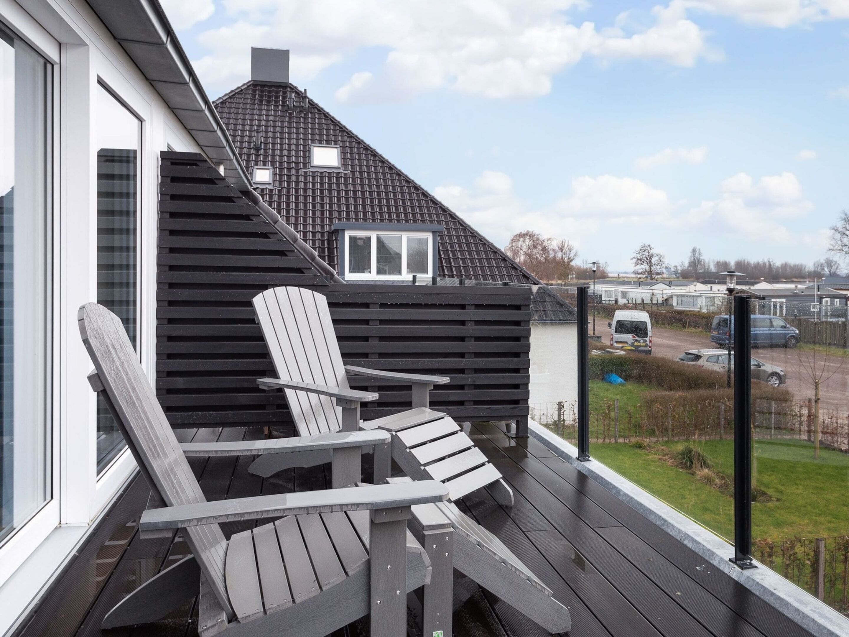 A deck with two Adirondack chairs, a house with a sloped roof, and a view of a road and parked cars in the background.