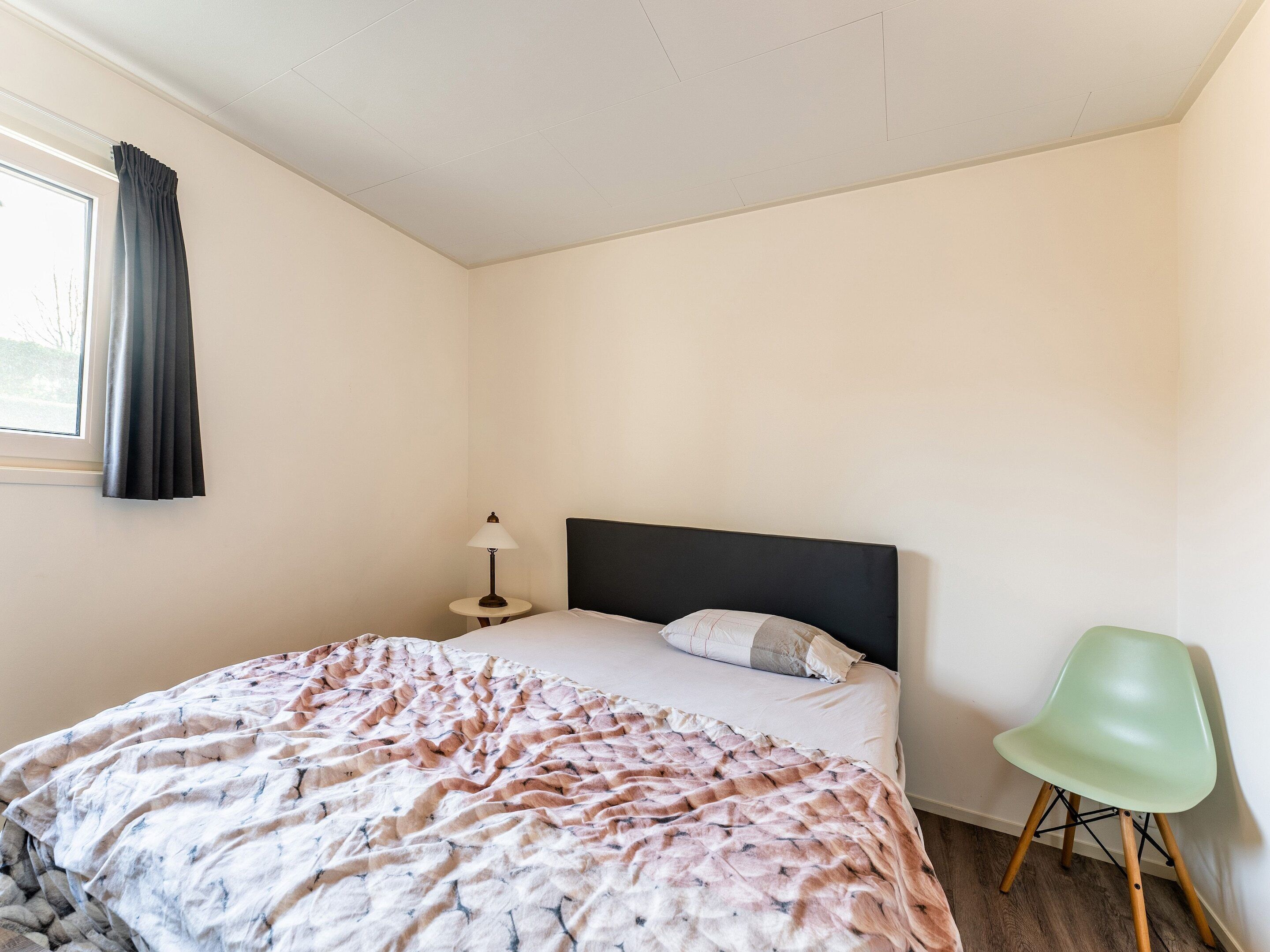 A bedroom with a bed, a nightstand with a lamp, a green chair, and a window with curtains.