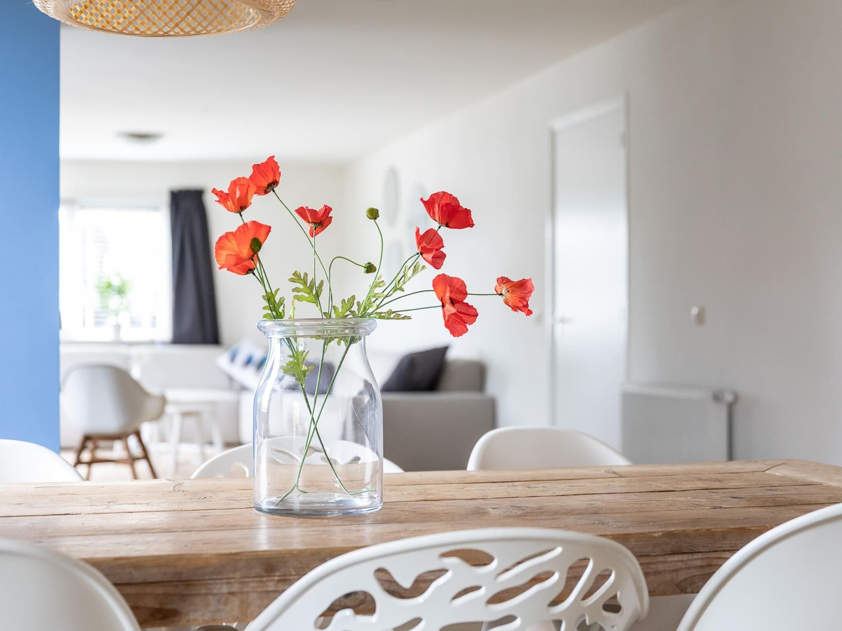 Een eettafel met een glazen vaas met rode bloemen, witte stoelen en een houten tafelblad.