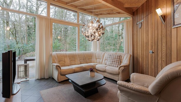 A cozy living room with a sofa, a coffee table, and a TV. Large windows offer a view of the forest.