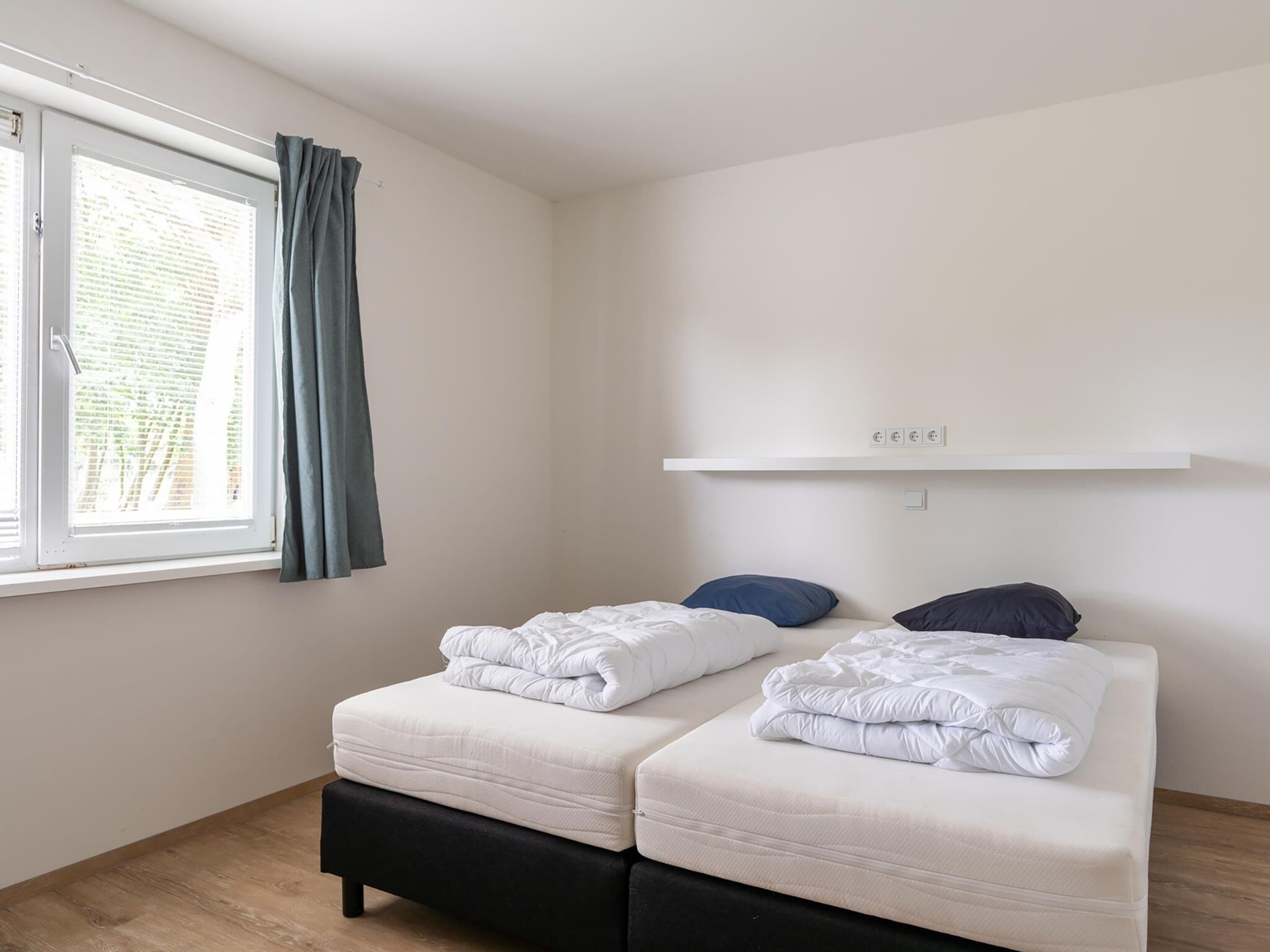 Una habitación pequeña y limpia con dos camas, una ventana con cortinas y una pared blanca con una estantería sobre las camas.