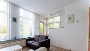 A modern living room with a sofa, a white table, and a radiator. There are large windows with blinds, a picture on the wall, and a door. - Waterside Stay With Private Jetty (Heeg)