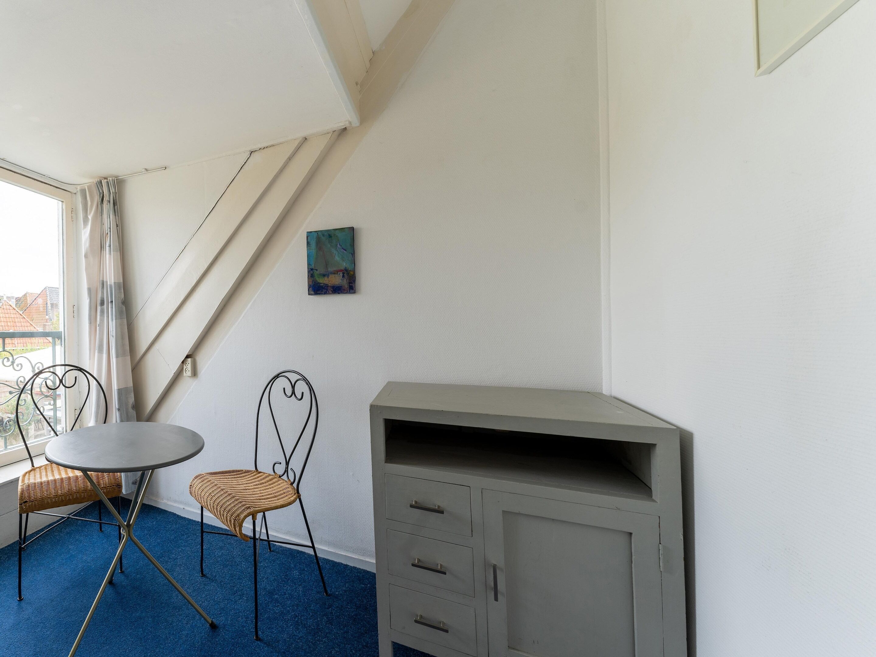 A room with a blue carpet, two wicker chairs, a small round table, a cabinet, and a painting on the wall.