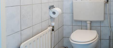 Une petite salle de bain propre, équipée de toilettes, d’un radiateur et d’un porte-rouleau de papier toilette.