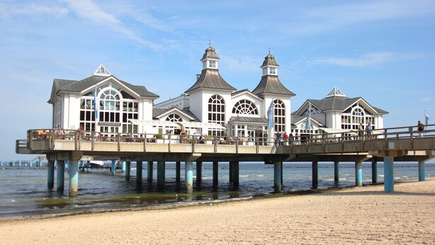 Eine Steg, der sich über den Strand erstreckt, mit einem weißen Gebäude, das mehrere Rundbogenfenster und eine zentrale Kuppel aufweist.
