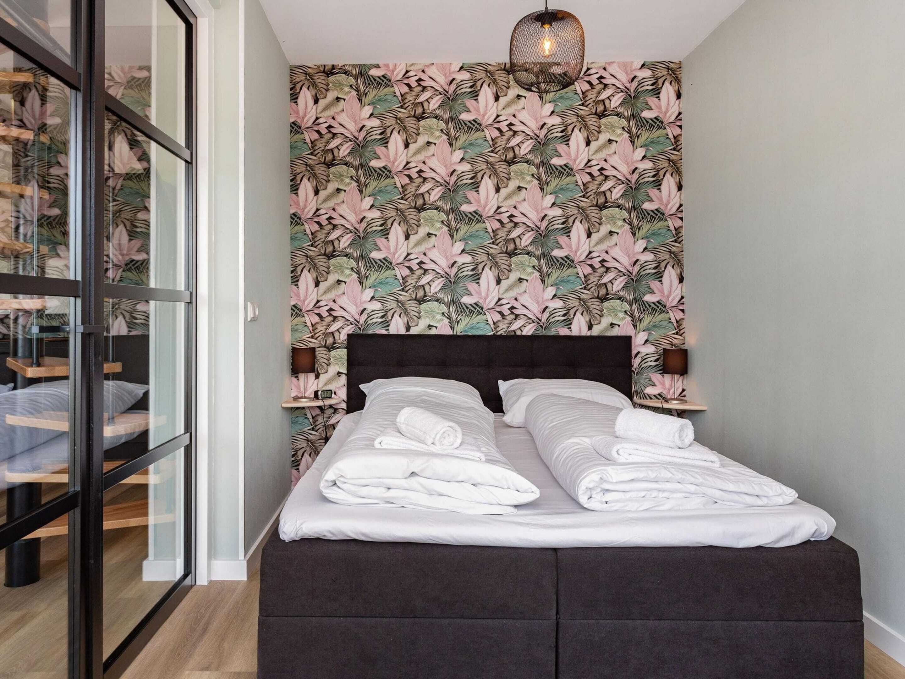 A bed with white linens and a dark headboard, a wall with a tropical leaf pattern, and a hanging light fixture.