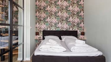 A bed with white linens and a dark headboard, a wall with a tropical leaf pattern, and a hanging light fixture.