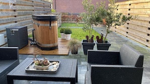 A patio with wicker furniture, a wooden barrel, potted plants, and a brick building in the background. - Holiday Home Paesens Near Wadden Sea (Paesens)