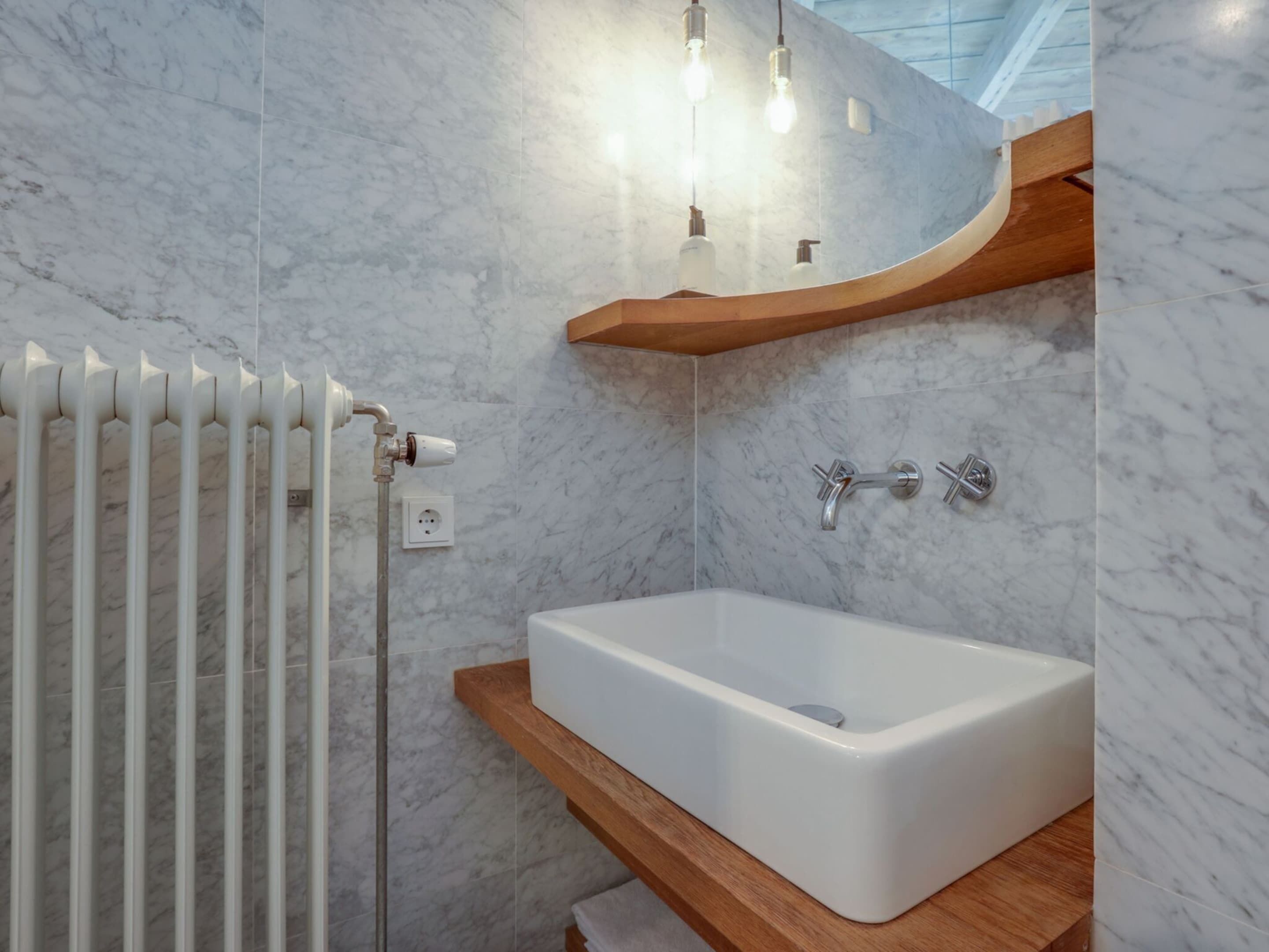 A modern bathroom with a white bathtub, marble walls, and a wooden shelf above the sink.