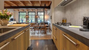 Una cocina moderna con armarios de madera, encimera negra y estufa integrada. Hay una zona de comedor con sillas de madera y una mesa, y ventanas amplias que dejan entrar luz natural.