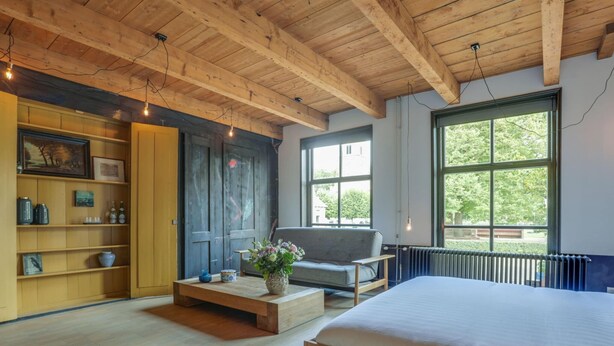 Un dormitorio moderno con techo de madera, una cama grande, un sofá, una mesa de centro y una ventana con vista a follaje.