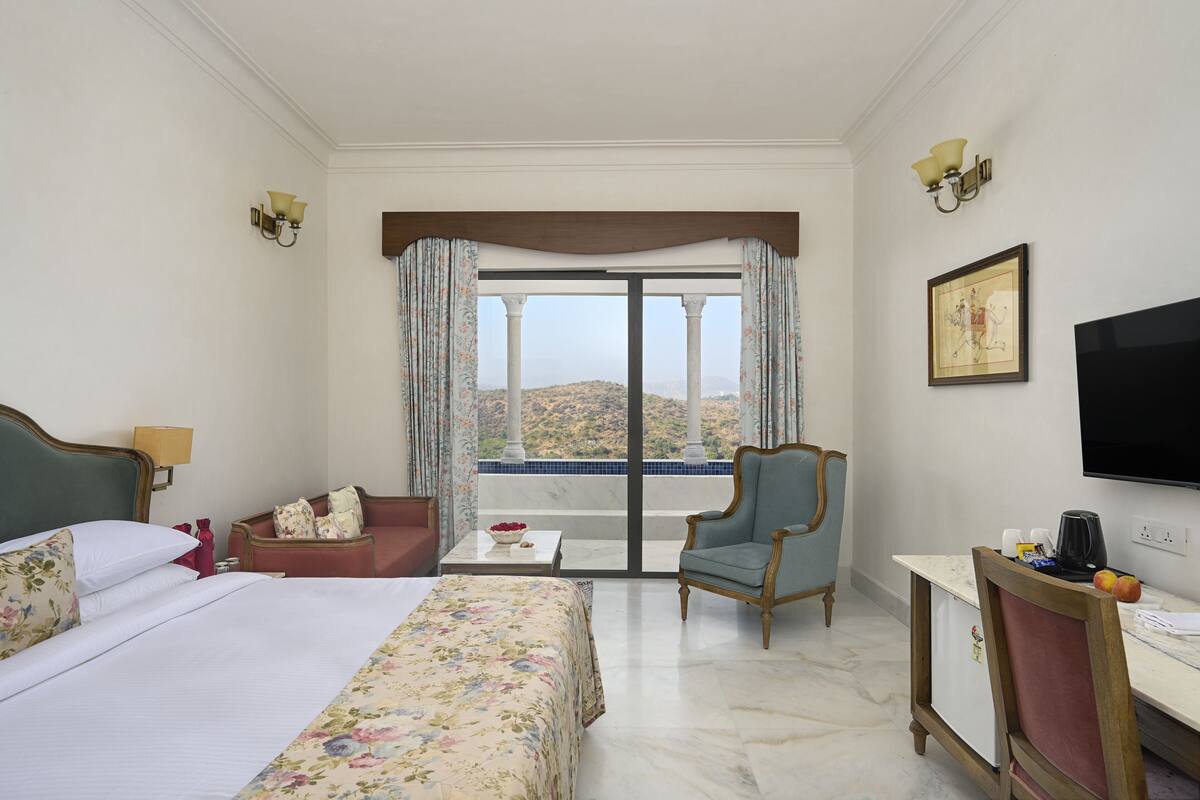 A hotel room with a bed, a desk, a chair, a TV, and a balcony with a view of the hills.