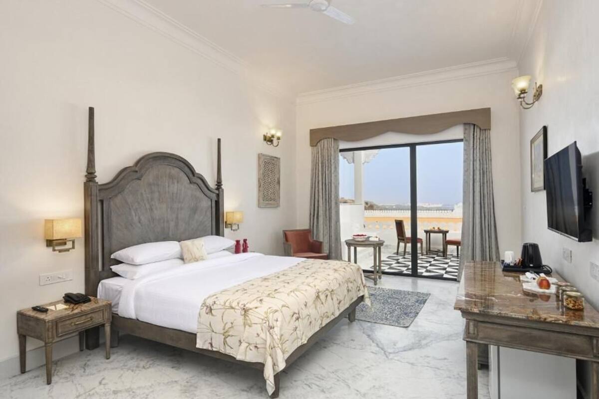 A hotel room with a large bed, a desk, a chair, a TV, and a balcony with a view.