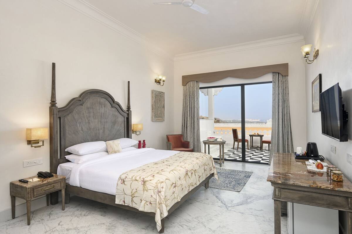 A hotel room with a large bed, a desk, a chair, a TV, and a balcony with a view.