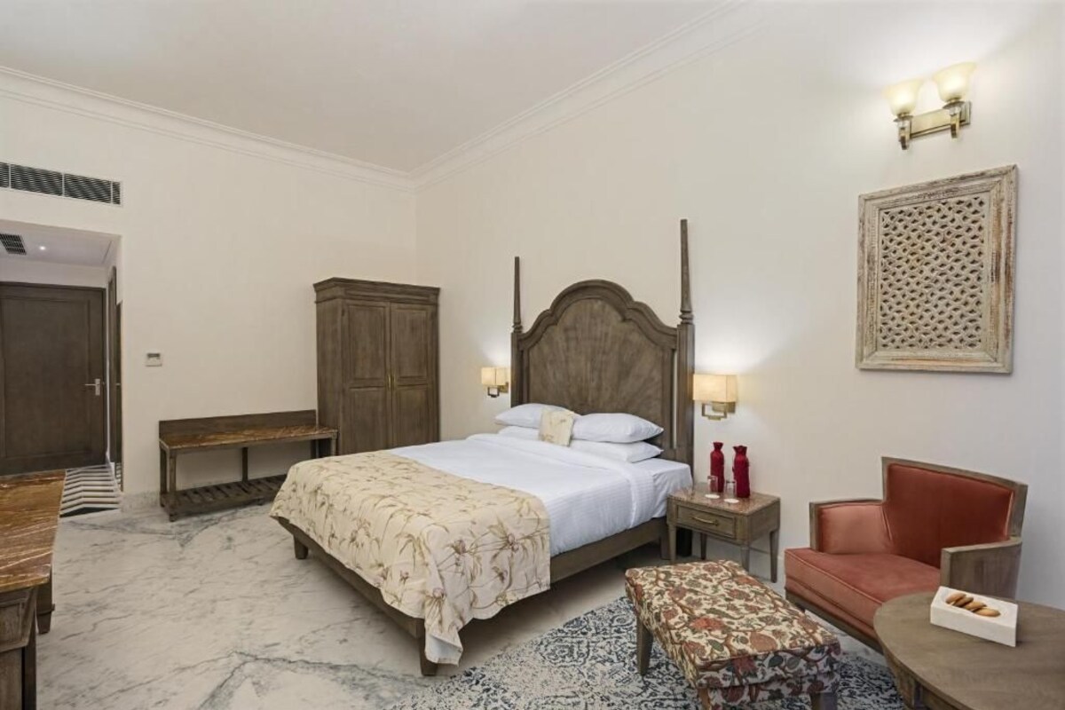 A bedroom with a four-poster bed, a wooden bench, a red chair, a small table with a box of cookies, and a patterned rug.
