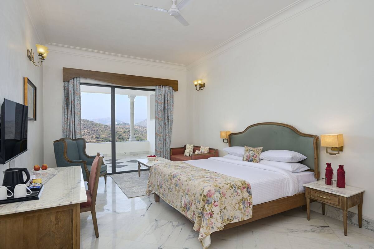 A hotel room with a bed, a desk with a coffee maker, a chair, a TV, and a view of the outdoors.