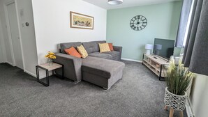 Ein Wohnzimmer mit grauem Ecksofa, einem kleinen Beistelltisch mit Blumen in einer Vase, einer Uhr an der Wand und einer Pflanze in einem weißen Topf.