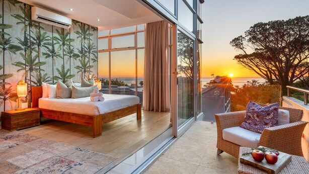 Ein Schlafzimmer mit einem großen Bett, einem Nachttisch, einer Lampe und Blick auf den Sonnenuntergang durch große Fenster.