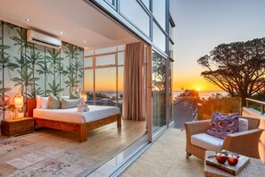 Un dormitorio con una cama grande, una mesita de noche, una lámpara y vistas a la puesta de sol a través de amplias ventanas.