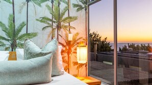Ein Schlafzimmer mit großem Fenster, einem Bett mit Kissen, einem Nachttisch, einer Lampe und Blick auf Palmen und einen Sonnenuntergang.