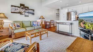Amplia sala de estar con sofá, mesa de centro y muebles de mimbre. Se ve una cocina con armarios blancos y un comedor con sillas.