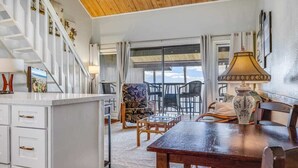 Un espacio amplio con techo de madera, una mesa de comedor con sillas, una zona de cocina con un mueble blanco y una escalera con barandillas blancas.