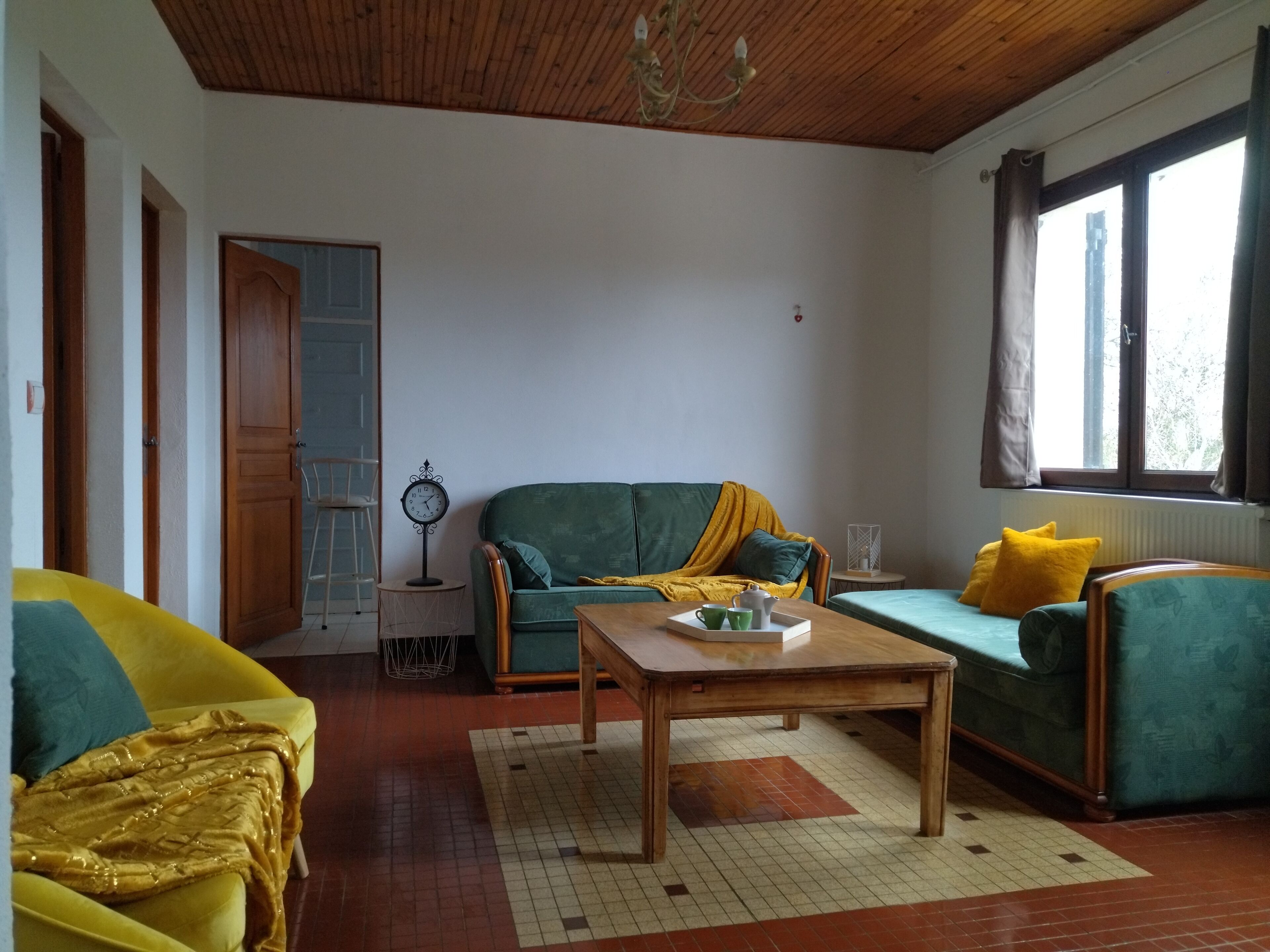 Un salón con techo de madera, un sofá verde con cojines amarillos, un sofá amarillo, una mesa de centro de madera y una ventana con cortinas.