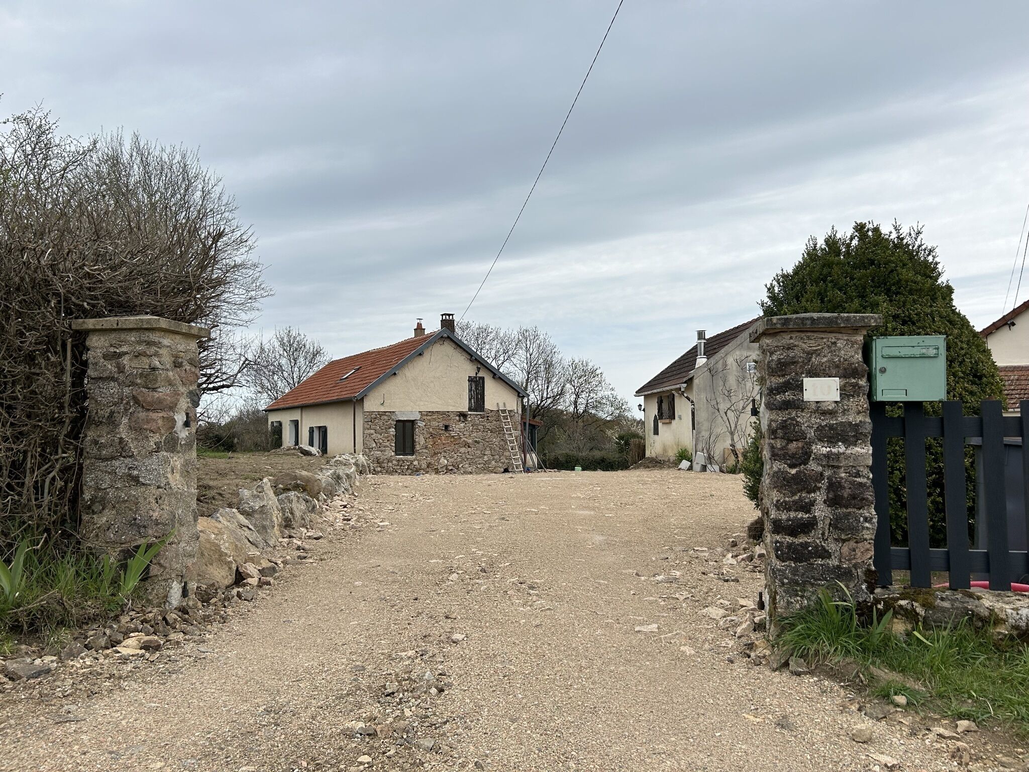 A rural property with a gravel driveway leading to a house with a stone wall and a metal gate.