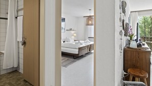 Habitación de hotel con puerta de madera, cama con sábanas blancas, mesita de noche con lámpara y vistas al exterior.