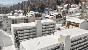 A modern multi-story building with a flat roof, likely a hotel or apartment complex, surrounded by snow-covered trees and a mountainous backdrop. - Nr.43 Haus Sotmirana by Interhome (Lenzerheide)