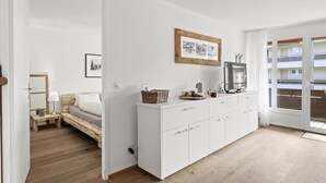 A modern bedroom with a bed, bedside table, and a dresser. There is a framed picture on the wall and a window with a view of buildings. - Nr.43 Haus Sotmirana by Interhome (Lenzerheide)