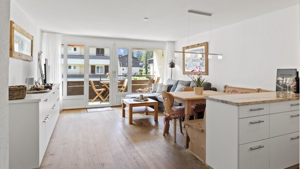 A modern kitchen with wooden flooring, a dining area with a table and chairs, and a view of a building through large glass doors. - Nr.43 Haus Sotmirana by Interhome (Lenzerheide)