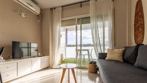Un soggiorno moderno con un divano grigio, un tavolino rotondo e una televisione su un mobile bianco. Si intravede un balcone con vista sull'acqua attraverso tende leggere.