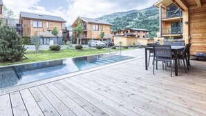 A modern outdoor pool area with wooden decking, a dining set, and a view of residential buildings in a mountainous area. - #5b mit Sauna & Pool by Interhome (Sankt Georgen am Kreischberg)