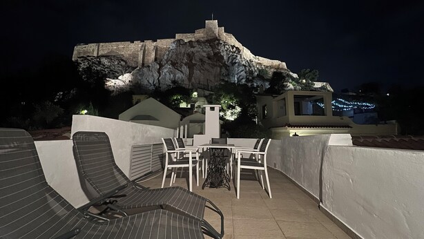 An outdoor seating area with lounge chairs and a table, overlooking a large rock formation and a building with lights. - In the shadow of Acropolis - Breathtaking view  (ATHENS)