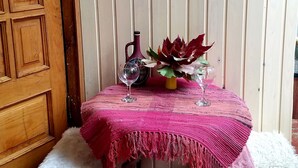Uma pequena mesa com toalha rosa, um vaso com flores vermelhas e duas taças de vinho. Ao fundo, uma porta de madeira e uma parede branca.
